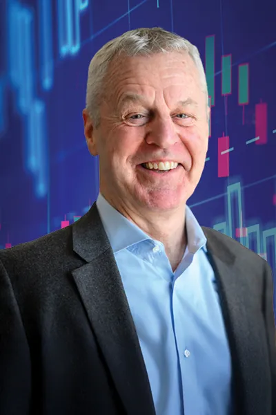 Smiling older man with short gray hair wearing a gray blazer and light blue shirt, set against a deep blue background overlaid with colorful financial candlestick chart graphics in teal, pink, and red.